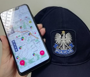 Policjant trzyma telefon w ręce z otwartą aplikacją Krajowa Mapa Zagrożeń a obok na biurku lez czapka policyjna